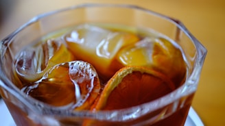 A close-up of a glass filled with iced tea, containing slices of citrus fruit and large ice cubes. The glass has a textured exterior, and the drink is a rich amber color.