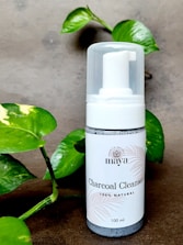 A bottle of charcoal cleanser with a white label and a pump dispenser, set against a neutral background. The product is surrounded by green leafy plants, adding a natural touch to the scene.