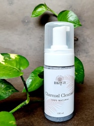 A bottle of charcoal cleanser with a white label and a pump dispenser, set against a neutral background. The product is surrounded by green leafy plants, adding a natural touch to the scene.