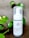 A bottle of charcoal cleanser with a white label and a pump dispenser, set against a neutral background. The product is surrounded by green leafy plants, adding a natural touch to the scene.