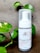 A bottle of charcoal cleanser with a white label and a pump dispenser, set against a neutral background. The product is surrounded by green leafy plants, adding a natural touch to the scene.