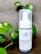 A bottle of charcoal cleanser with a white label and a pump dispenser, set against a neutral background. The product is surrounded by green leafy plants, adding a natural touch to the scene.
