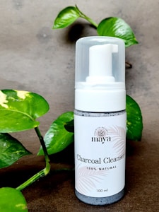 A bottle of charcoal cleanser with a white label and a pump dispenser, set against a neutral background. The product is surrounded by green leafy plants, adding a natural touch to the scene.