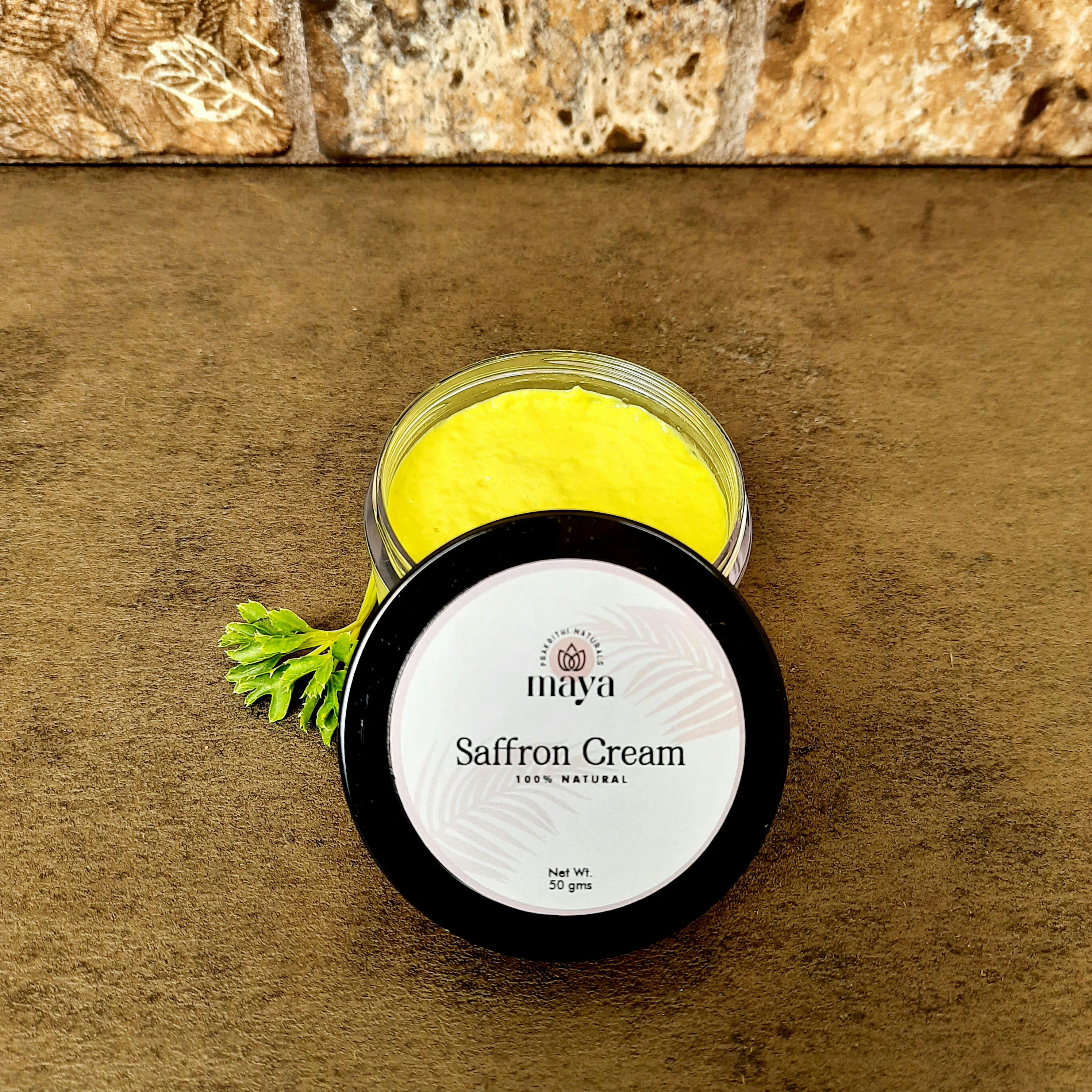 A jar of saffron cream with a bright yellow color is placed on a brown textured surface. The lid is off to the side and displays the label 'maya, Saffron Cream, 100% Natural, Net Wt. 50 gms.' A small green leaf is nearby, adding a natural element. The background features a stone wall with a rough texture.