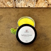 A jar of saffron cream with a bright yellow color is placed on a brown textured surface. The lid is off to the side and displays the label 'maya, Saffron Cream, 100% Natural, Net Wt. 50 gms.' A small green leaf is nearby, adding a natural element. The background features a stone wall with a rough texture.