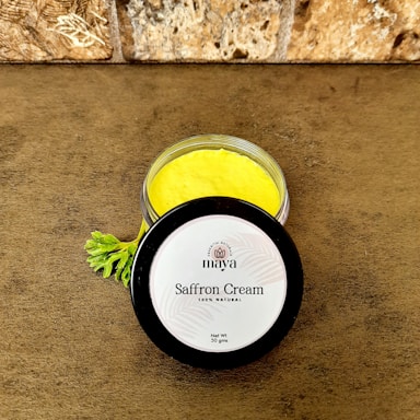A jar of saffron cream with a bright yellow color is placed on a brown textured surface. The lid is off to the side and displays the label 'maya, Saffron Cream, 100% Natural, Net Wt. 50 gms.' A small green leaf is nearby, adding a natural element. The background features a stone wall with a rough texture.