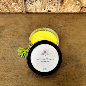 A jar of saffron cream with a bright yellow color is placed on a brown textured surface. The lid is off to the side and displays the label 'maya, Saffron Cream, 100% Natural, Net Wt. 50 gms.' A small green leaf is nearby, adding a natural element. The background features a stone wall with a rough texture.