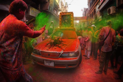 A vibrant street scene with people celebrating, covered in colorful powders. A decorated car with flowers is in the center, surrounded by a crowd engaging in festivities. The air is filled with green and orange powders as participants throw them enthusiastically. Buildings line the narrow street, forming a lively atmosphere.