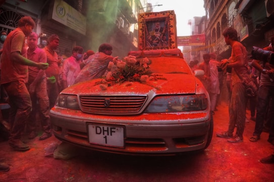 A street scene filled with people covered in colorful powders, likely celebrating a festival. A car in the foreground is adorned with flowers and a framed picture, also covered in vibrant powders. The street is bustling with activity, and the atmosphere is energetic and lively.