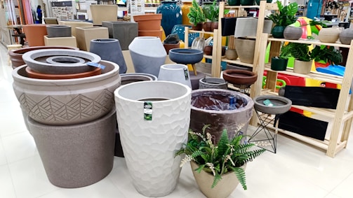 Various large planters and pots are displayed in a store setting, arranged neatly on shelves and the floor. They come in diverse shapes, sizes, and textures, some with decorative patterns. The space is bright and organized, with plants inside some pots.