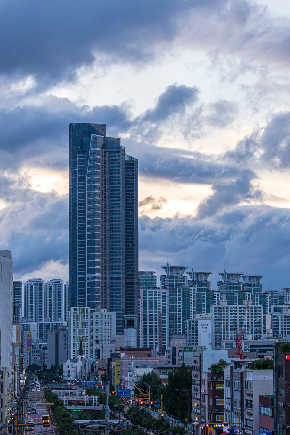 The image depicts a cityscape at dusk, with a large skyscraper standing tall against an overcast sky. The building's facade is illuminated by warm orange and yellow lights, creating a striking contrast to the cool blue hues of the sky. The city street below is lined with various buildings, including one on each side of the frame, showcasing the urban landscape. Cars are parked along the street, adding a sense of activity to the scene.