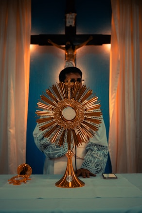 An ornate gold monstrance sits on a table covered with a white cloth, with a religious figure in robes standing behind it. In the background, a crucifix is mounted on a blue wall, illuminated by warm lighting from the sides.