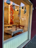 A cozy restaurant setting is visible through a large window. Inside, tables are set with checkered cloths, plates, glassware, and folded napkins. Soft lighting and warm tones create an inviting atmosphere. Several decorative cloths hang from a line, adding a rustic charm.