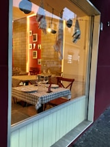 A cozy restaurant setting is visible through a large window. Inside, tables are set with checkered cloths, plates, glassware, and folded napkins. Soft lighting and warm tones create an inviting atmosphere. Several decorative cloths hang from a line, adding a rustic charm.