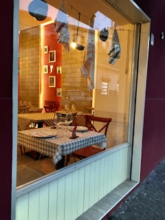 A cozy restaurant setting is visible through a large window. Inside, tables are set with checkered cloths, plates, glassware, and folded napkins. Soft lighting and warm tones create an inviting atmosphere. Several decorative cloths hang from a line, adding a rustic charm.