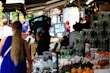 A busy marketplace scene with vendors and customers interacting. A person wearing gloves carries a crate on their shoulder. Various fresh produce such as grapes and oranges are displayed on stalls. People are visible in the background, some standing and others walking past.
