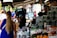 A busy marketplace scene with vendors and customers interacting. A person wearing gloves carries a crate on their shoulder. Various fresh produce such as grapes and oranges are displayed on stalls. People are visible in the background, some standing and others walking past.