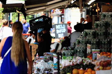 A busy marketplace scene with vendors and customers interacting. A person wearing gloves carries a crate on their shoulder. Various fresh produce such as grapes and oranges are displayed on stalls. People are visible in the background, some standing and others walking past.