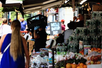 A busy marketplace scene with vendors and customers interacting. A person wearing gloves carries a crate on their shoulder. Various fresh produce such as grapes and oranges are displayed on stalls. People are visible in the background, some standing and others walking past.