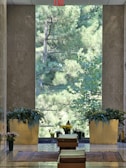 A serene interior space with a large window that showcases a lush, green view of trees outside. The room includes two large golden planters filled with vibrant greenery and flowers. There are small statues and more flowers placed near a small step or ledge. The floor features a polished stone surface with decorative rugs adding warmth to the space. An 'Exit' sign is visible at the top of the window structure.