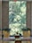 A serene interior space with a large window that showcases a lush, green view of trees outside. The room includes two large golden planters filled with vibrant greenery and flowers. There are small statues and more flowers placed near a small step or ledge. The floor features a polished stone surface with decorative rugs adding warmth to the space. An 'Exit' sign is visible at the top of the window structure.