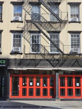 A multi-story building with beige walls and black window frames. There are multiple air conditioning units installed in the windows. The building features a fire escape with metal stairs and railings, casting detailed shadows on the facade. The ground floor has bright red doors and windows that appear to be part of a commercial establishment.