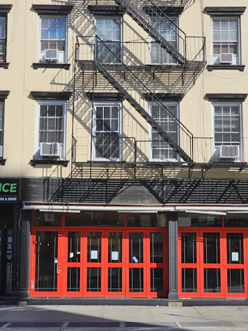 A multi-story building with beige walls and black window frames. There are multiple air conditioning units installed in the windows. The building features a fire escape with metal stairs and railings, casting detailed shadows on the facade. The ground floor has bright red doors and windows that appear to be part of a commercial establishment.