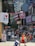 A bustling city street scene with numerous billboards and signs visible, including an 'I Love NY' sign and various advertisements. Several people are walking on the sidewalk, some carrying shopping bags. A man in the foreground is pulling a cooler on wheels. The background showcases tall buildings with more signs and digital displays.