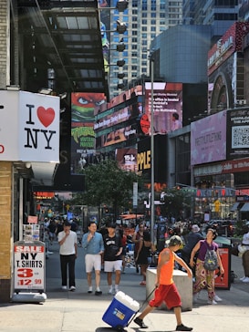 A bustling city street scene with numerous billboards and signs visible, including an 'I Love NY' sign and various advertisements. Several people are walking on the sidewalk, some carrying shopping bags. A man in the foreground is pulling a cooler on wheels. The background showcases tall buildings with more signs and digital displays.