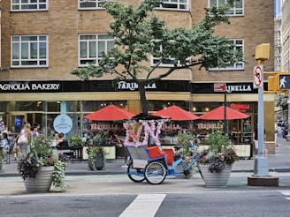 A busy city street corner features a charming bakery and pizzeria with large windows in a brick building. Red umbrellas provide shade for outdoor seating, and there are large flower pots with colorful plants along the sidewalk. A blue pedicab adorned with pink floral decorations is parked nearby, creating a whimsical touch. People are walking along the sidewalk, adding to the bustling urban atmosphere.
