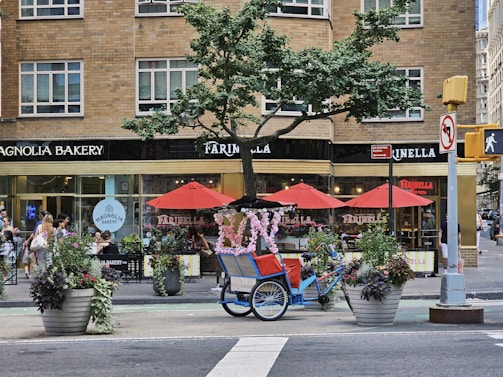 A busy city street corner features a charming bakery and pizzeria with large windows in a brick building. Red umbrellas provide shade for outdoor seating, and there are large flower pots with colorful plants along the sidewalk. A blue pedicab adorned with pink floral decorations is parked nearby, creating a whimsical touch. People are walking along the sidewalk, adding to the bustling urban atmosphere.