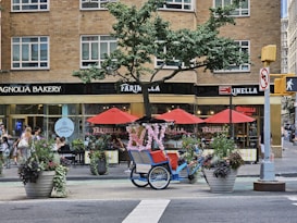 A busy city street corner features a charming bakery and pizzeria with large windows in a brick building. Red umbrellas provide shade for outdoor seating, and there are large flower pots with colorful plants along the sidewalk. A blue pedicab adorned with pink floral decorations is parked nearby, creating a whimsical touch. People are walking along the sidewalk, adding to the bustling urban atmosphere.