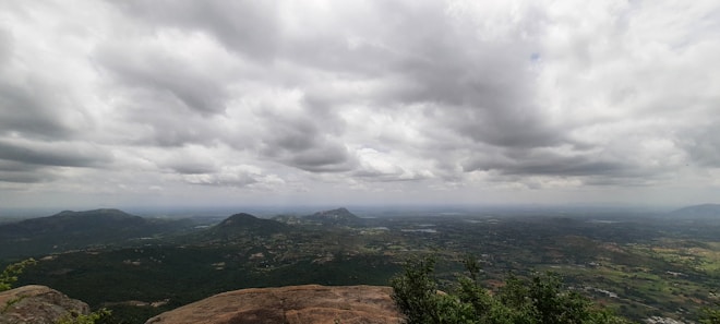 A panoramic view from a high vantage point overlooking a vast landscape with mountains, valleys, and scattered patches of greenery. The foreground includes rocky terrain and some bushes, while the sky is filled with large, dense clouds, giving it a dramatic appearance.