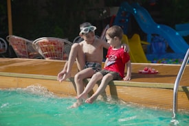 Two children are sitting on the edge of a pool, dipping their feet into the water. One child is wearing swim goggles on their forehead, while the other is wearing a red shirt. There are some colorful slides and chairs in the background.