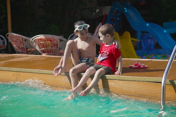 Two children are sitting on the edge of a pool, dipping their feet into the water. One child is wearing swim goggles on their forehead, while the other is wearing a red shirt. There are some colorful slides and chairs in the background.