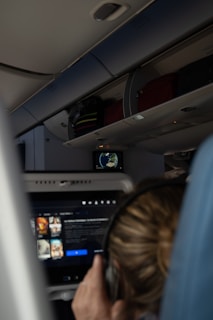 A person on an airplane is watching a screen on the seatback. The screen shows a movie selection interface, while in the background, the overhead compartments are partially visible along with another screen displaying a world map.