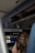 A person on an airplane is watching a screen on the seatback. The screen shows a movie selection interface, while in the background, the overhead compartments are partially visible along with another screen displaying a world map.