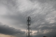 A tall telecommunications tower stands against a backdrop of dark, dramatic clouds, suggesting an impending storm. The sky is filled with layers of textured clouds, shading the scene in tones of gray and white.