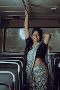 A woman wearing a patterned sari stands inside an empty bus. She gracefully holds onto the overhead railing with one hand while resting the other on her hip. Seats are visible in the background, and soft light creates a calm atmosphere.