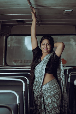 A woman wearing a patterned sari stands inside an empty bus. She gracefully holds onto the overhead railing with one hand while resting the other on her hip. Seats are visible in the background, and soft light creates a calm atmosphere.