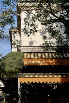 A building facade with a green and orange striped awning. There are trees in front with branches casting shadows on the wall. The words 'Les Blouses Blanches' are written on the awning.
