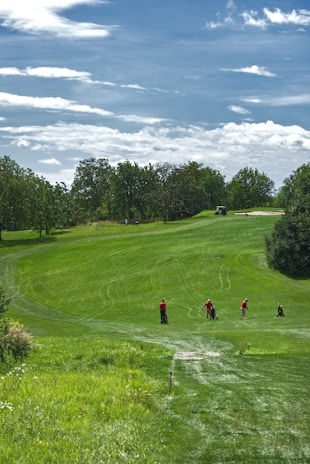 A lush green golf course with a rolling fairway under a partly cloudy sky. Four golfers are seen on the fairway, wearing red shirts, and are either walking or preparing to play. Trees surround the course, adding to the serene, natural setting.