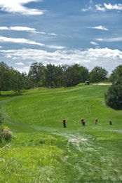A lush green golf course with a rolling fairway under a partly cloudy sky. Four golfers are seen on the fairway, wearing red shirts, and are either walking or preparing to play. Trees surround the course, adding to the serene, natural setting.