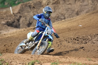 A motorcyclist wearing protective gear and a helmet competes in an off-road motocross race on a dirt track. The rider leans into a turn, kicking up dirt in an action-packed scene.