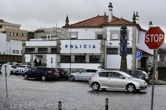 A street scene featuring a building marked with 'POLICIA' indicating a police station. Several parked cars are visible, including a police car with a checkered blue pattern. The street is paved with cobblestones, and there is a visible stop sign in the foreground. The sky is overcast, suggesting a gloomy atmosphere.