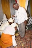 A couple dressed in traditional attire is kneeling on a decorative mat, while an older man stands before them, raising his hand in a gesture. The setting appears to be ceremonial, with vibrant decorations of red, yellow, and white drapery in the background.