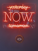 A neon sign with the words 'yesterday', 'NOW', and 'tomorrow', where 'yesterday' and 'tomorrow' are struck through, emphasizing the word 'NOW'. Below the sign hangs a decorative item featuring an eye surrounded by gold embellishments against a dark background.
