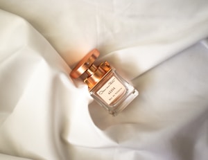 A bottle of perfume with a metallic cap lies on a soft, white fabric. The label on the bottle displays the word 'Alibi' along with additional text in smaller font. The overall ambiance is one of elegance and simplicity, enhanced by the gentle folds and clean texture of the fabric.