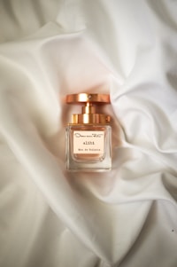 A perfume bottle with a gold cap sits on a soft, white fabric background. The label reads 'alibi Eau de Toilette' and features elegant typography. The lighting gives the scene a luxurious and sophisticated feel.