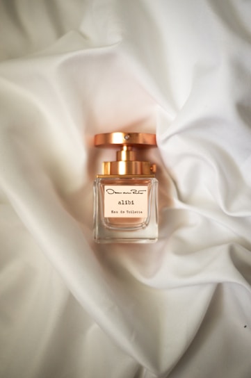 A perfume bottle with a gold cap sits on a soft, white fabric background. The label reads 'alibi Eau de Toilette' and features elegant typography. The lighting gives the scene a luxurious and sophisticated feel.